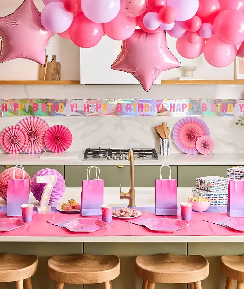 Pink birthday party table with a balloon arch, star balloons, a birthday banner, gift bags, party tableware, and a lit number 7 decoration. Pink birthday party table with a balloon arch, star balloons, a birthday banner, gift bags, party tableware, and a lit number 7 decoration.